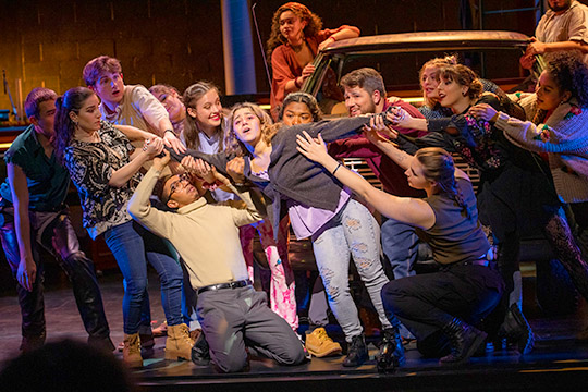 A large group of actors all surround a central young woman, seeming to pull her back and forth. A rusty old truck is behind them.